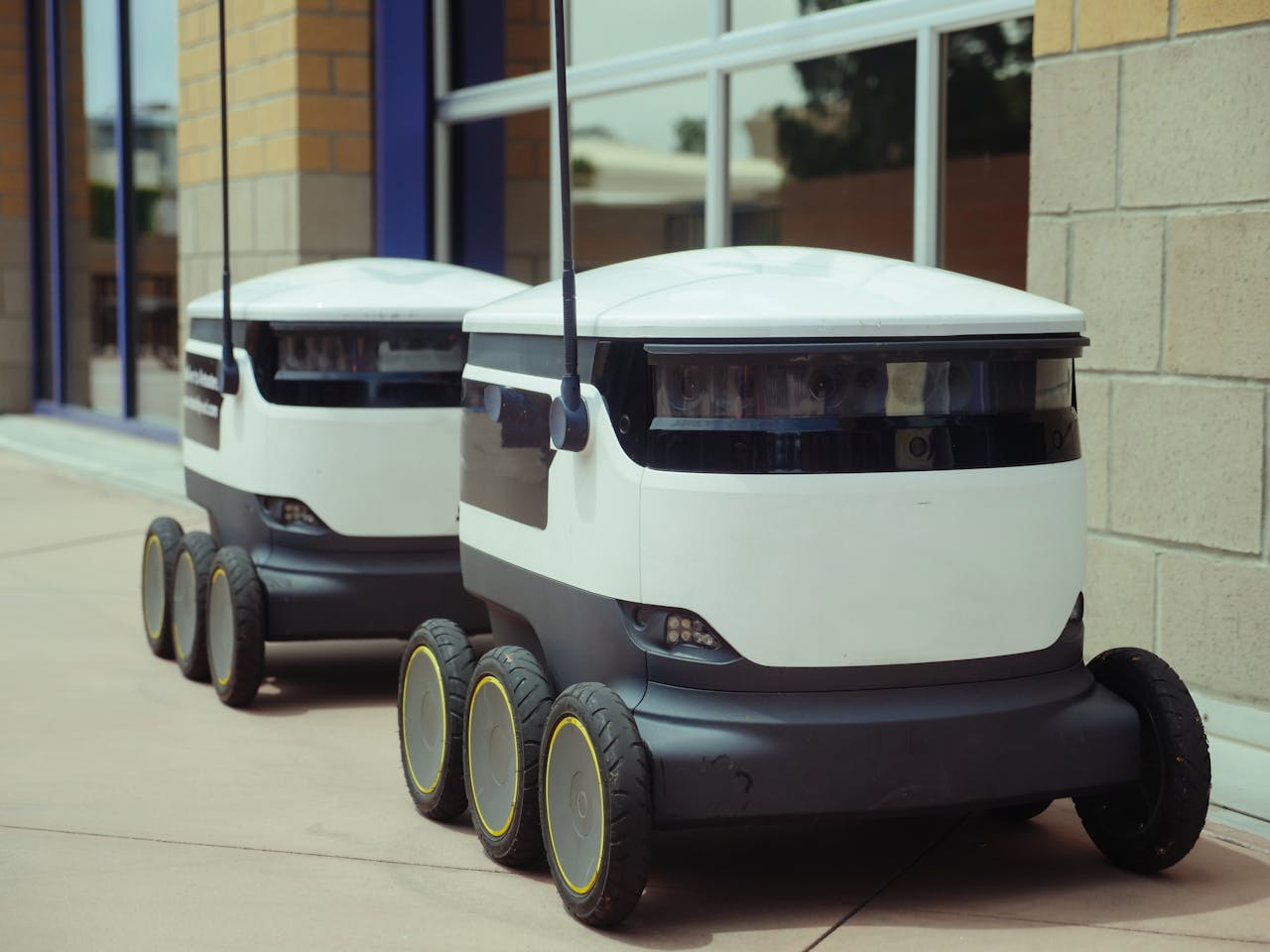 services-01 Two autonomous delivery robots positioned outside a modern building, showcasing innovation in robotics and mobility.